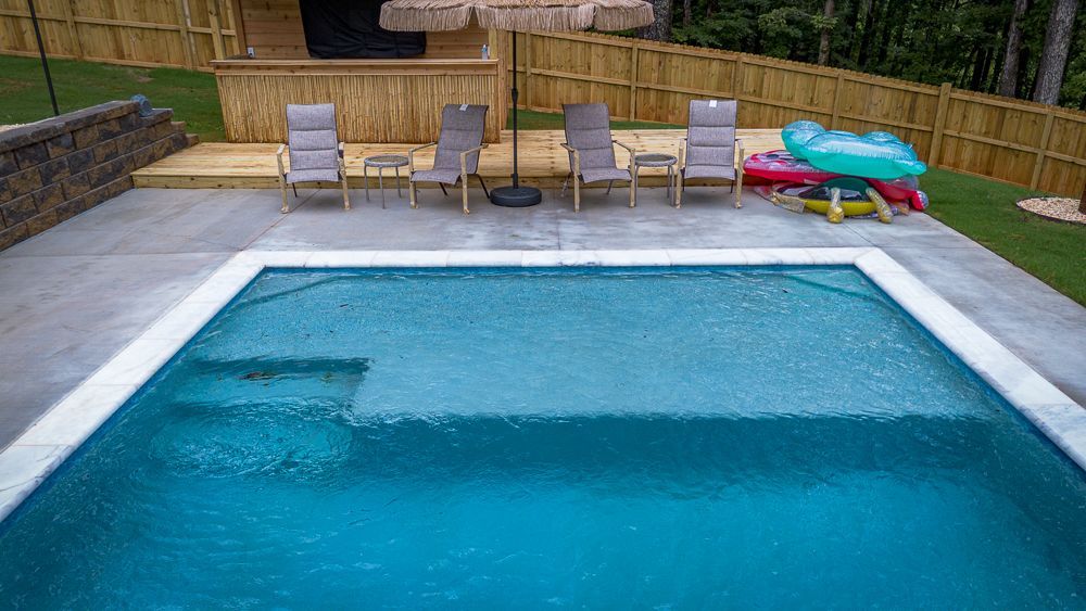 A backyard pool with chairs, umbrella, and inflatables, concrete patio, wooden fence and building in background.