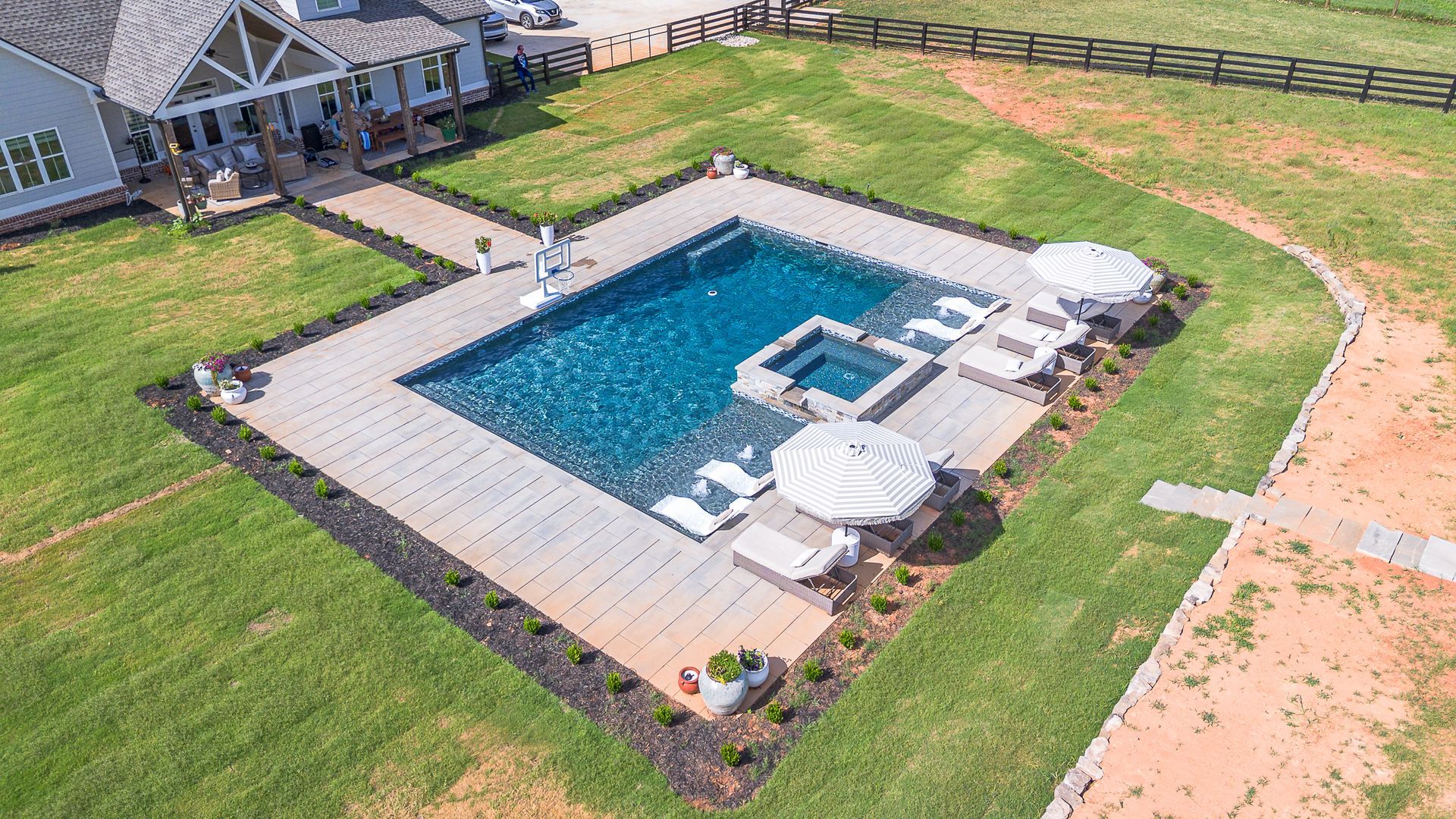 Aerial view of a backyard pool area with a gray patio, chairs, umbrellas, and a house in the background.