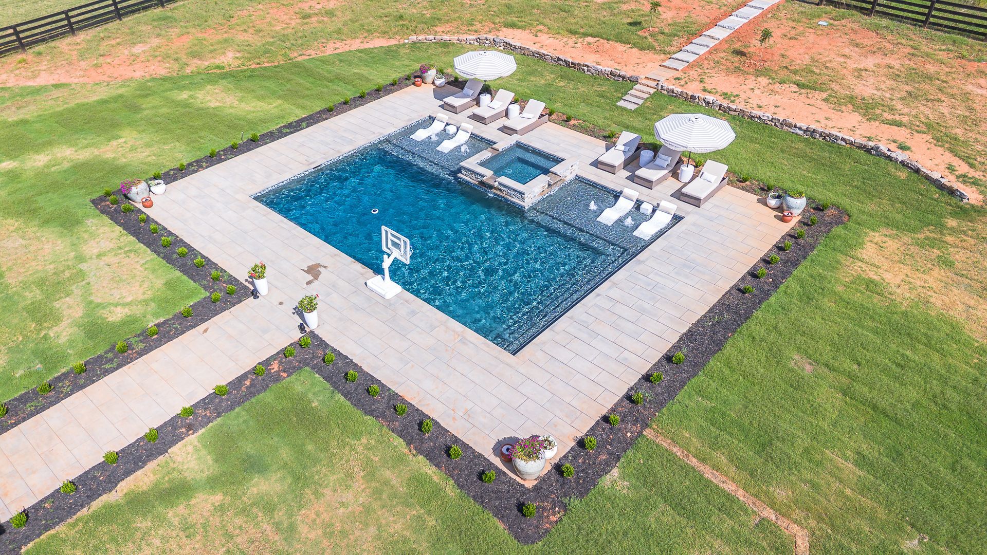 Aerial view of a luxurious pool area with lounge chairs, umbrellas, and a basketball hoop, surrounded by a grassy lawn.