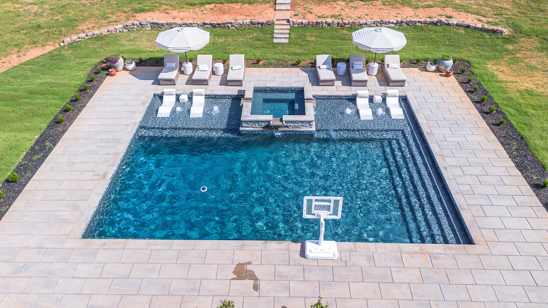 Outdoor pool with loungers, umbrellas, and a basketball hoop, surrounded by stone patio and lawn.