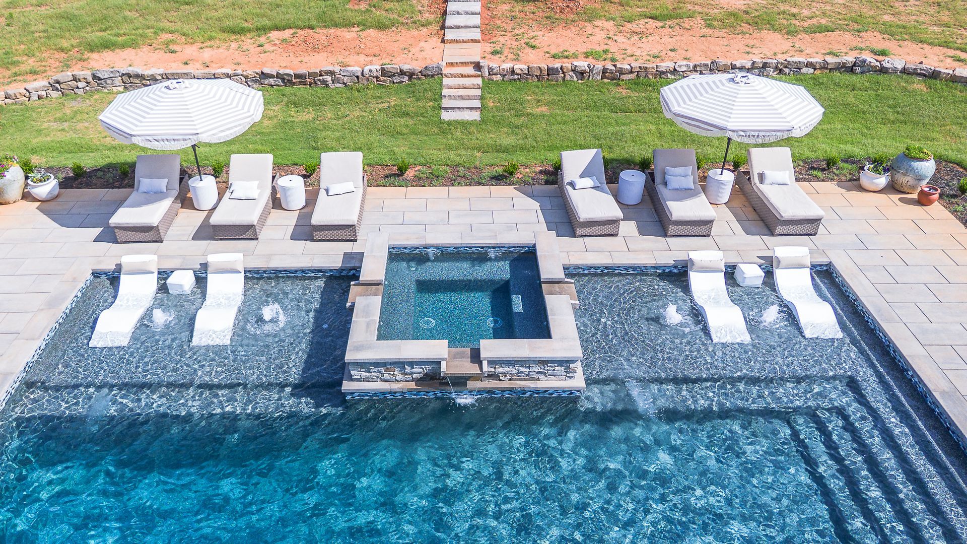 Luxury pool with loungers, umbrellas, and a spa area. Blue water, green grass, sunny day.