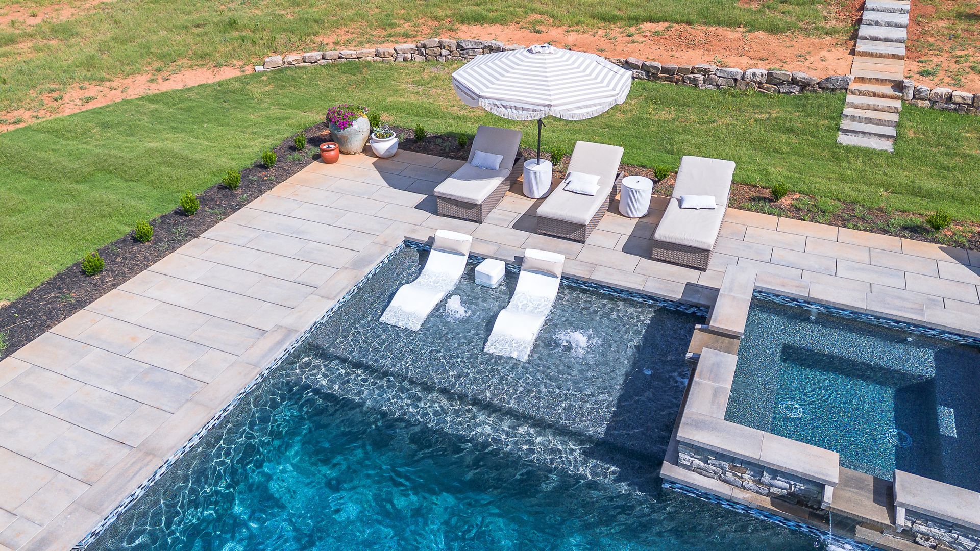 Overhead view of a pool with chaise lounges, a striped umbrella, and a hot tub, in a grassy setting.
