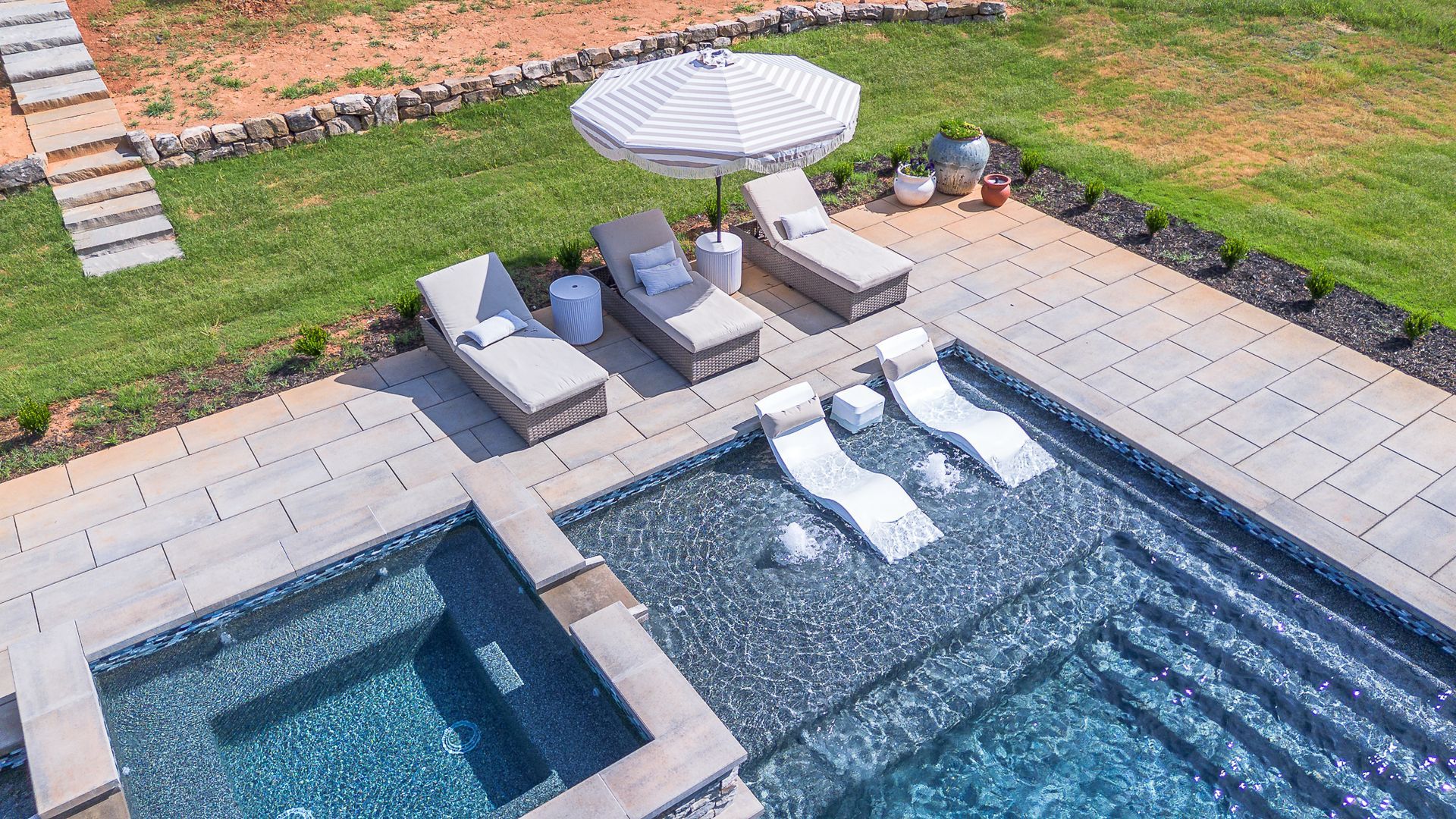 Pool area with lounge chairs, umbrella, and built-in seating in the water; grass and stonework surrounding.
