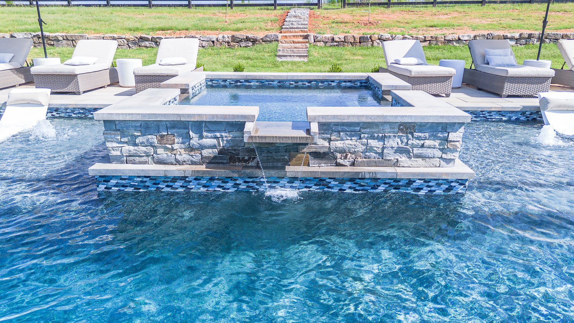 A pool with a raised spa feature, lounge chairs, and a grassy yard.