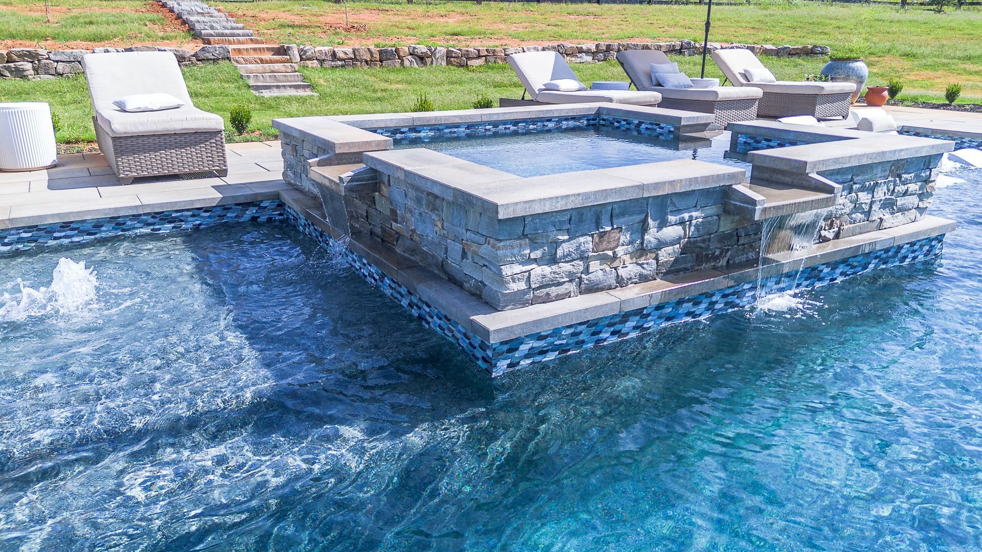 Pool with a spa, surrounded by stone and lounge chairs, steps in the background.