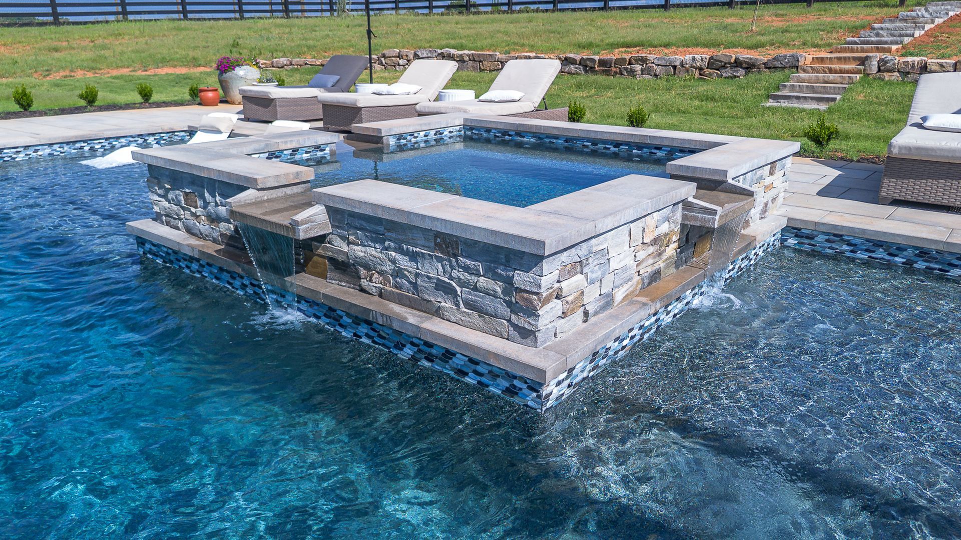 A luxurious pool with a built-in spa, stone accents, and cascading water features in a backyard setting.