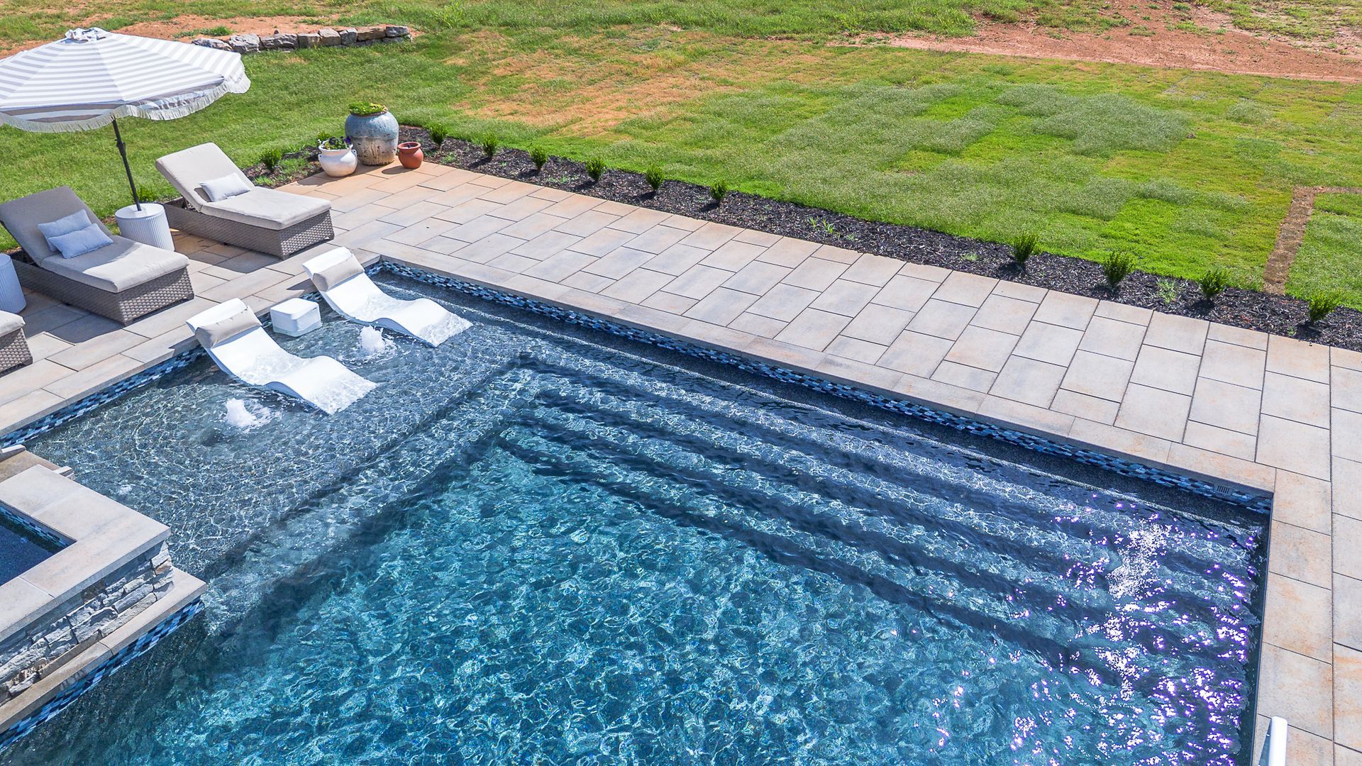 Pool with chaise lounges, umbrella, and steps. Bright blue water, gray patio, and green lawn.