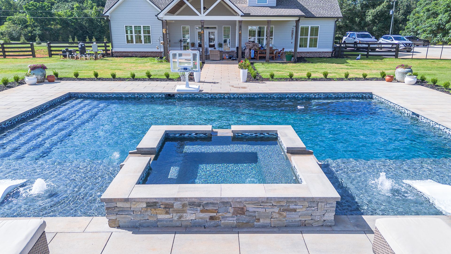 Luxury pool with a raised hot tub, in front of a large light blue house.