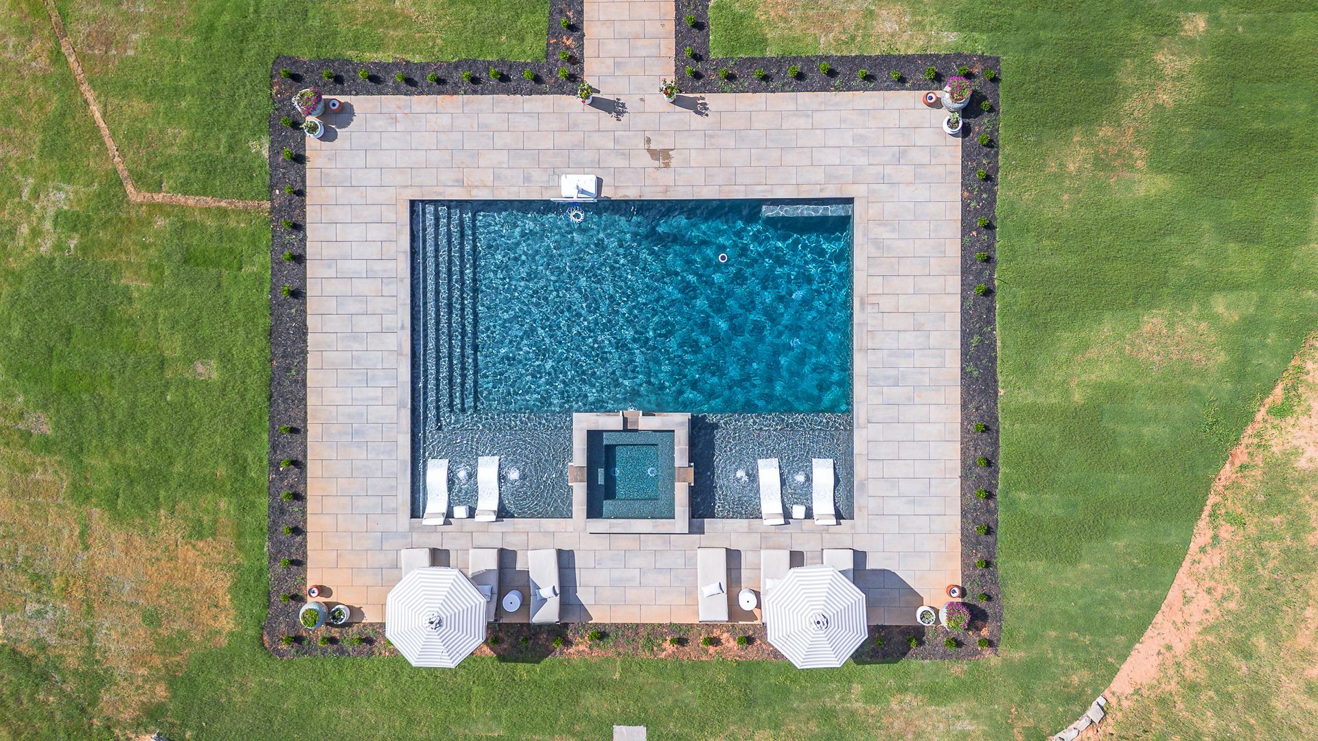 Overhead view of a square pool with lounge chairs and umbrellas surrounded by grass and stone.