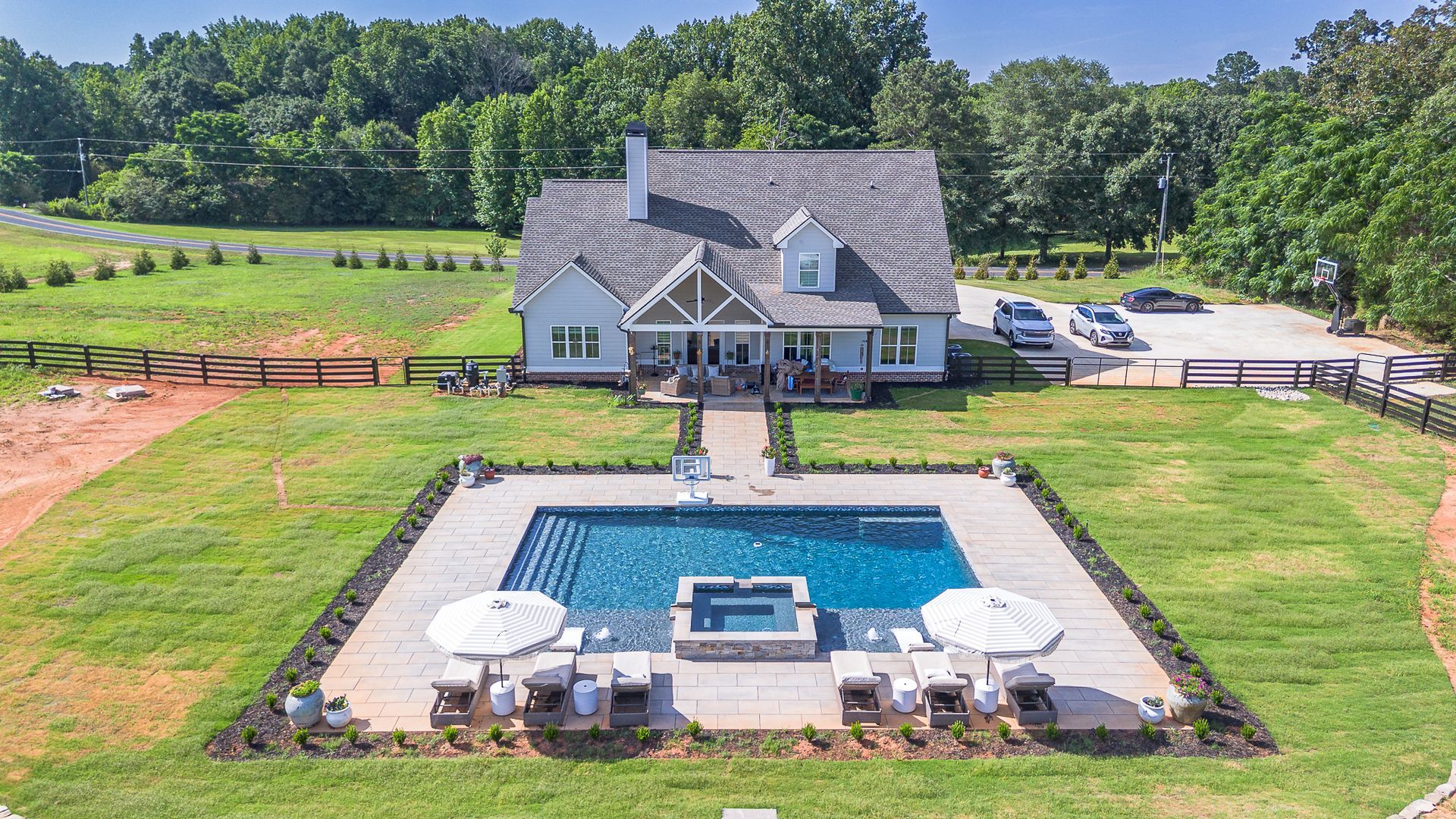 A house with a pool in front, surrounded by grass, with cars parked nearby and a field behind.