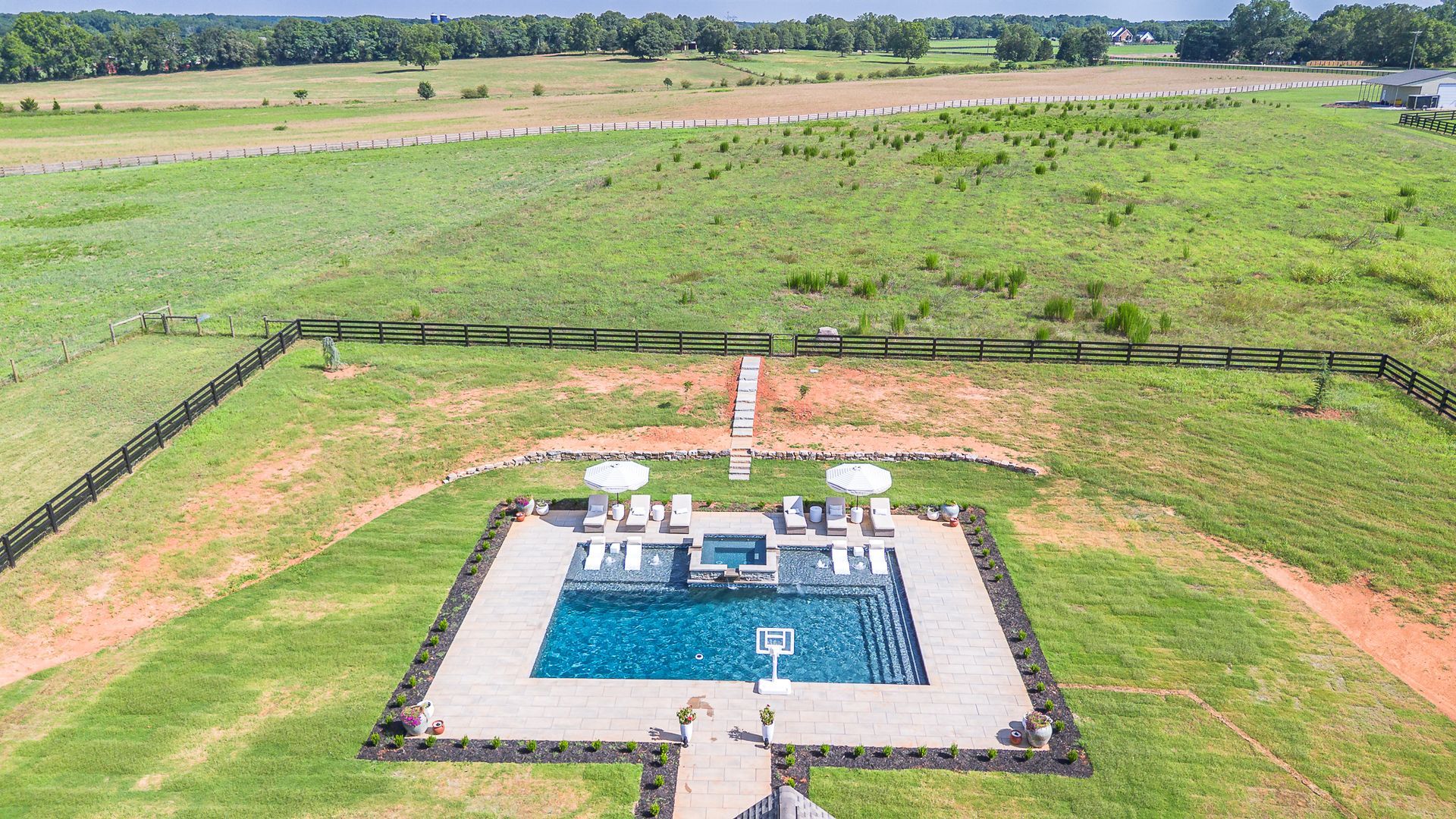 Aerial view of a backyard with a square pool, lounge chairs, and a vast green field beyond a black fence.