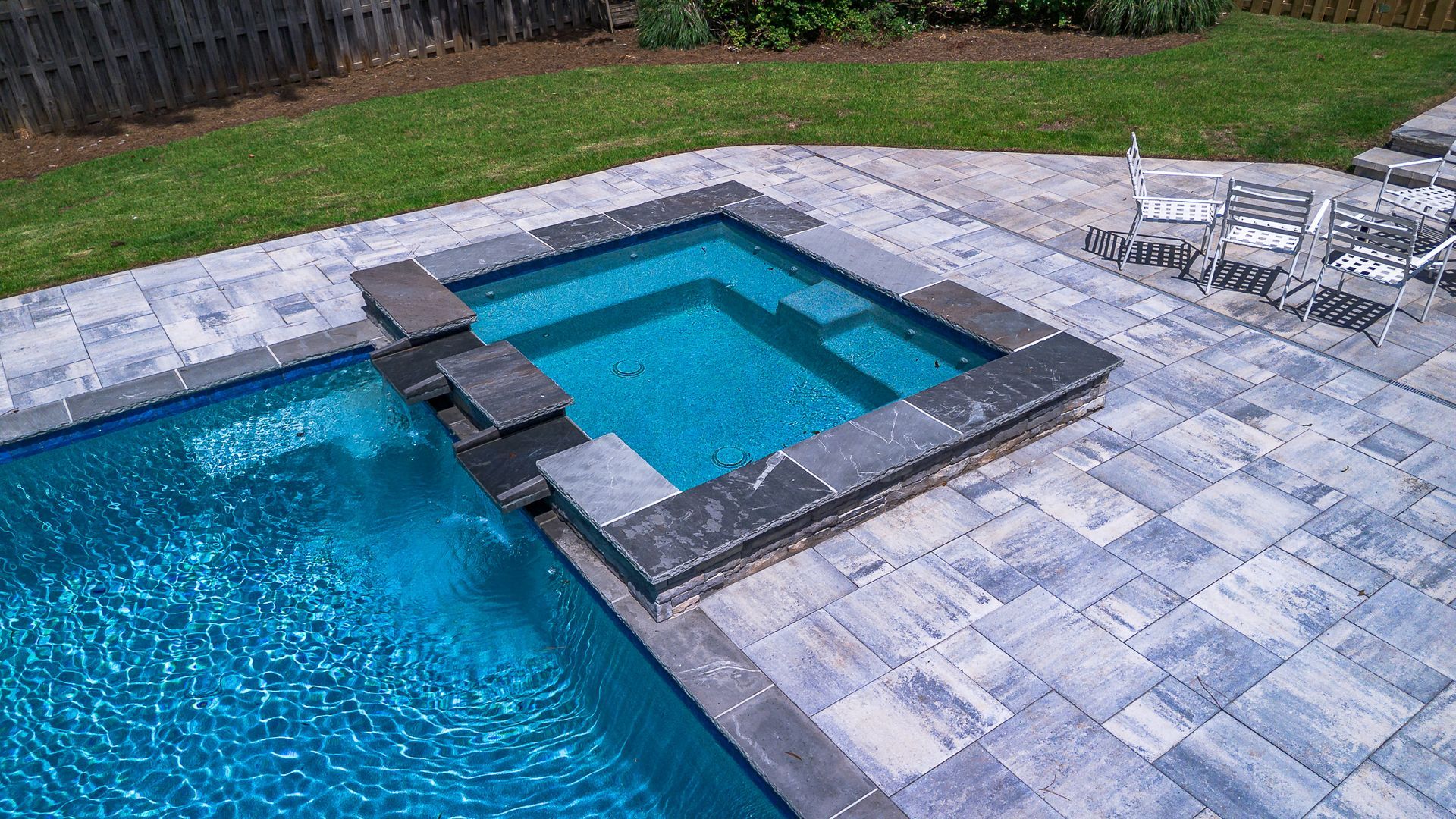 Pool with hot tub, blue water, gray pavers, green lawn.