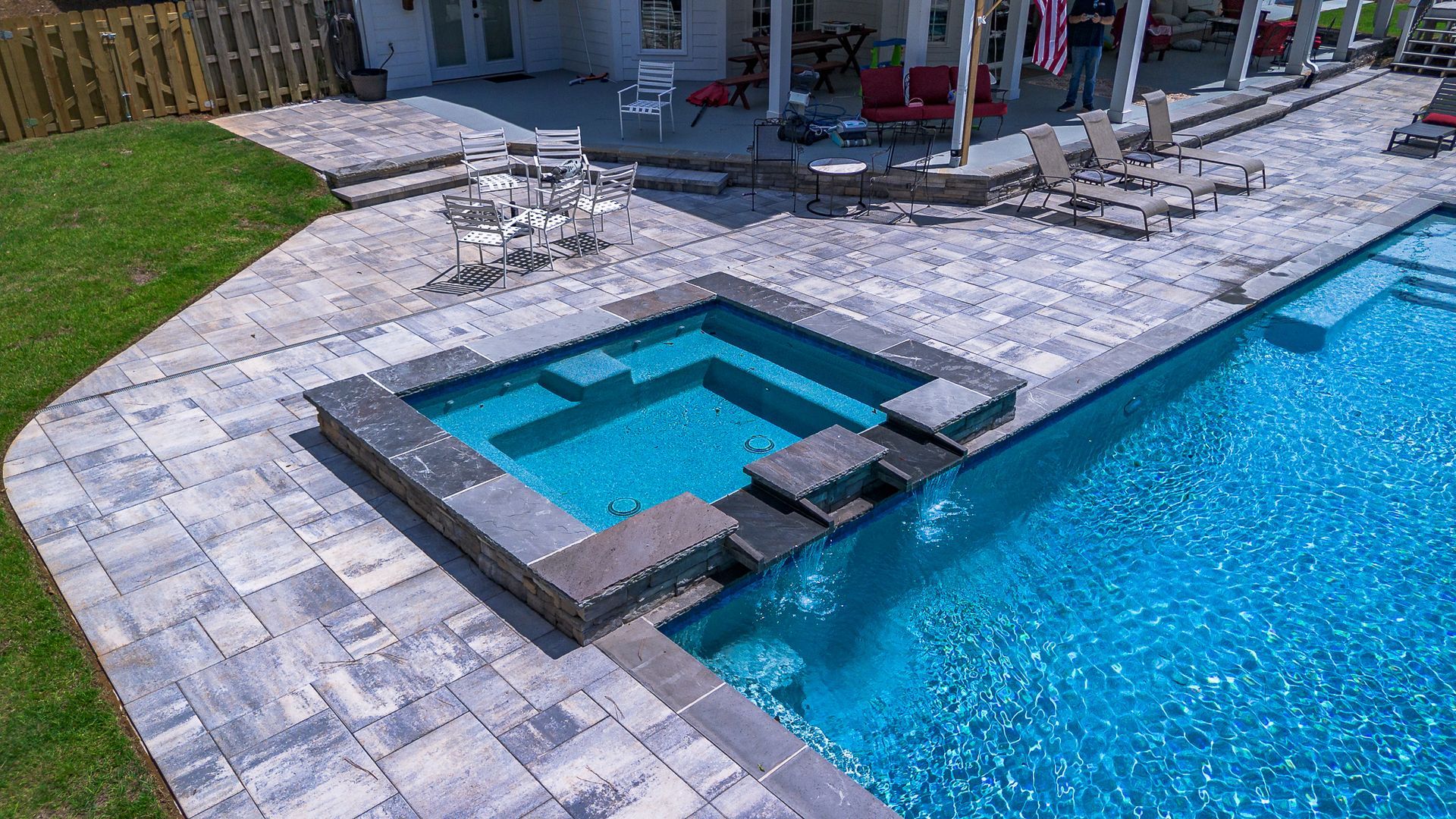 Stone patio with pool, spa, and steps to house. Turquoise water, gray pavers, green grass.