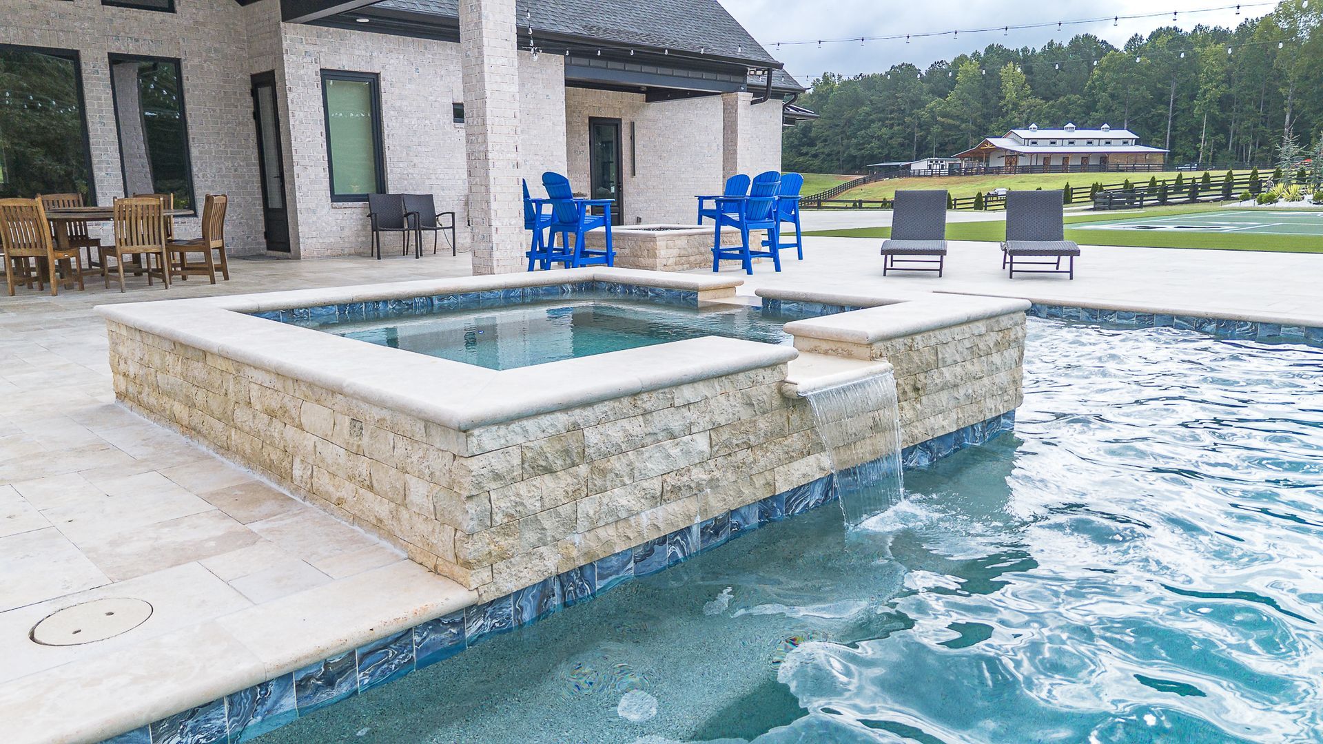 A luxury backyard pool with a spa, blue chairs, and a large house in the background.