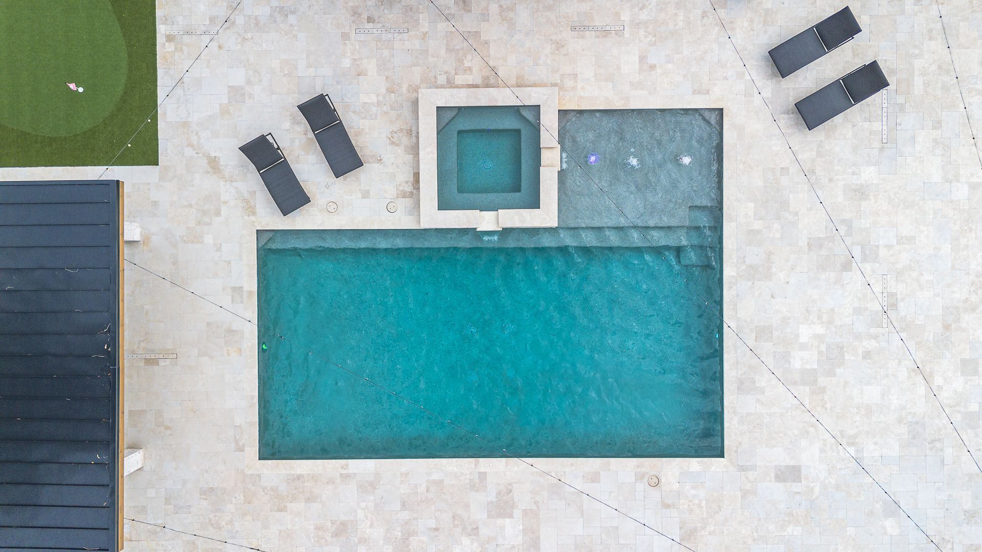 Overhead view of a swimming pool, hot tub, and putting green. Turquoise water surrounded by light-colored stone.