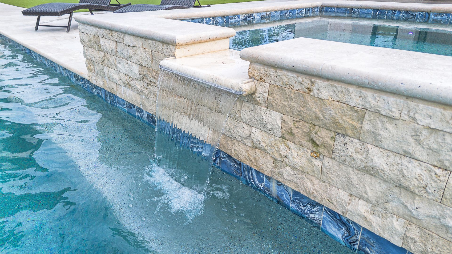 Pool edge with travertine stone, water cascading over, blue water, sunlit.