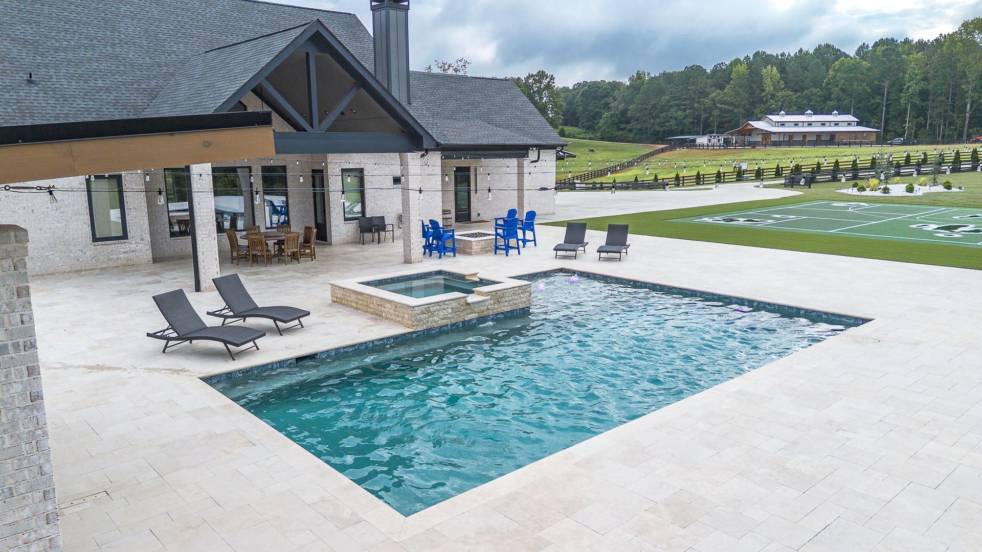 Backyard with a pool, jacuzzi, and lounge chairs, next to a light brick house with a dark roof.