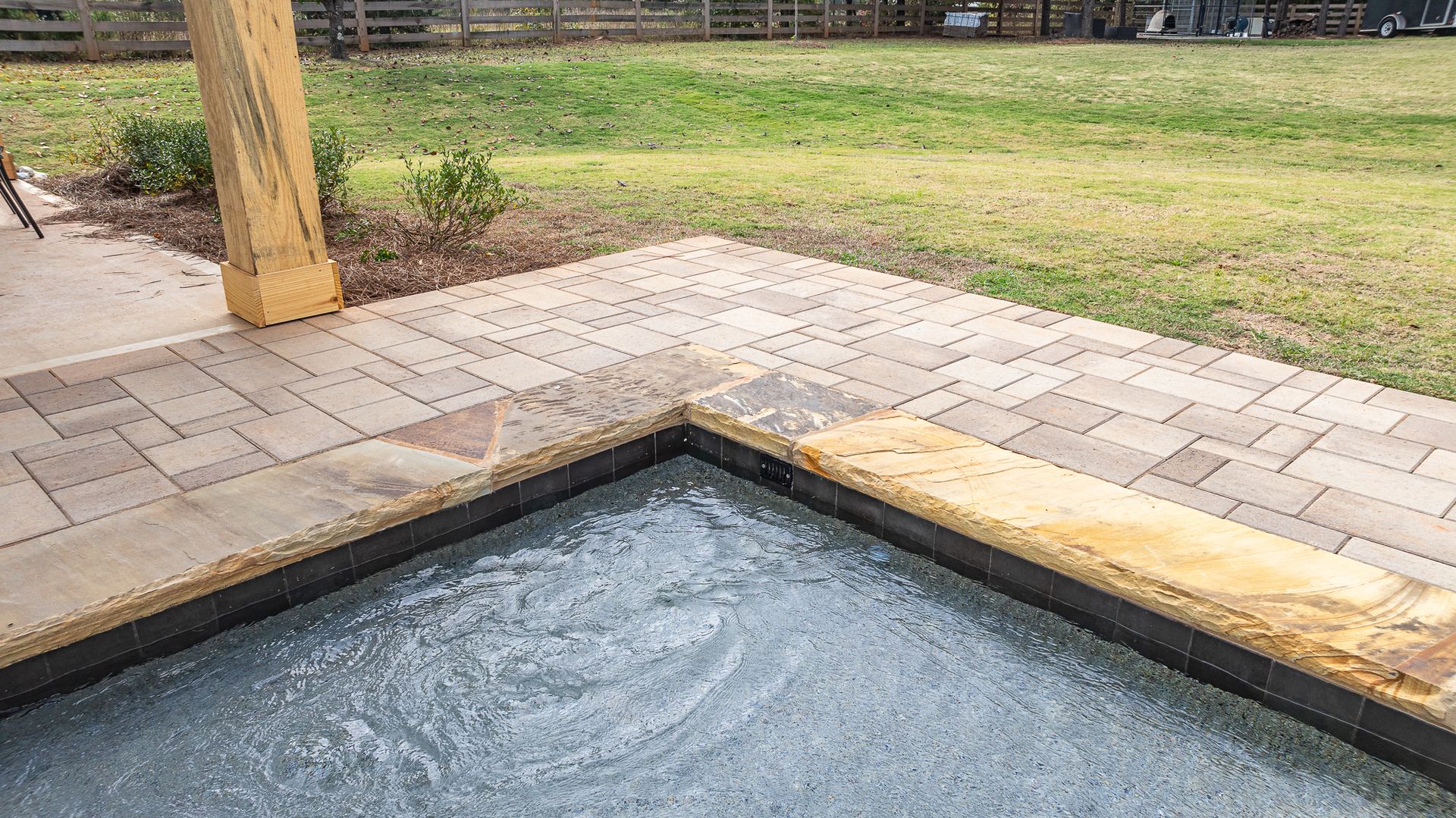 A swimming pool is surrounded by a brick patio and a lush green field.