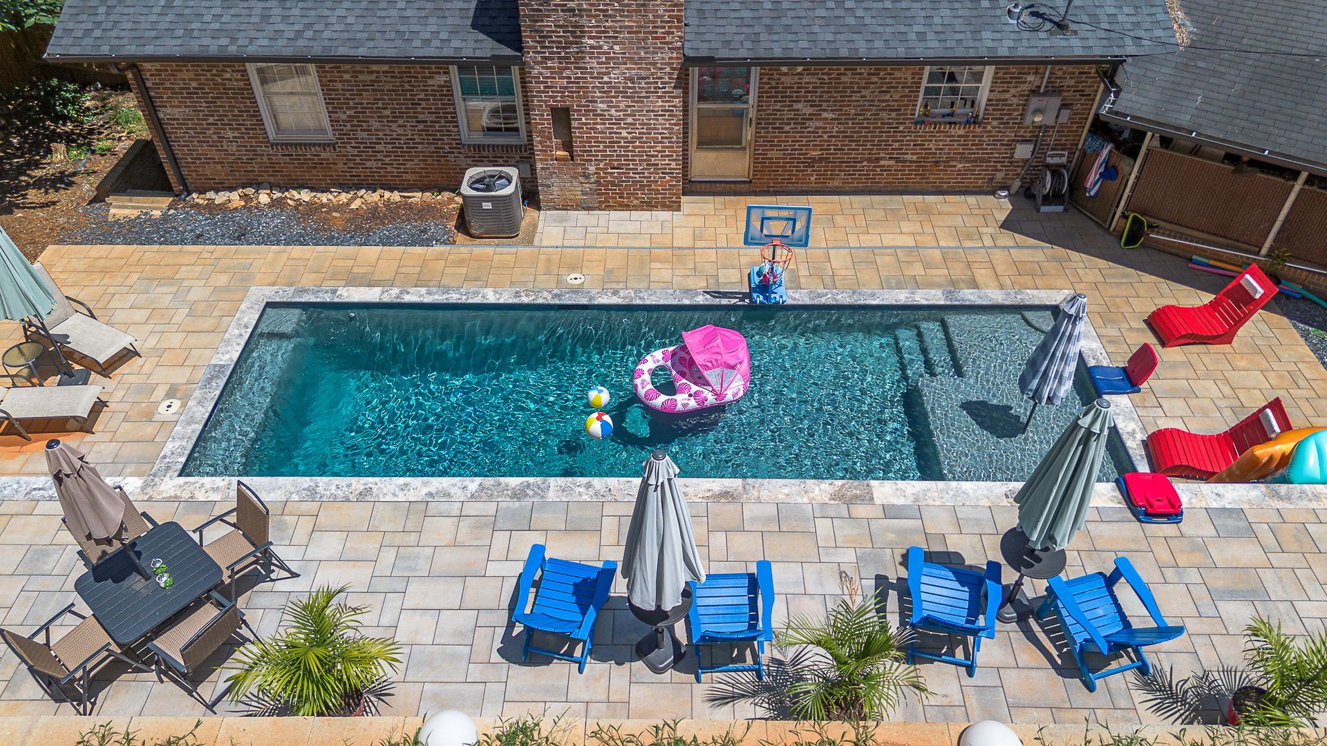 Overhead view of a backyard pool with blue chairs, lounge chairs, and a floating raft; a brick house is in the background.