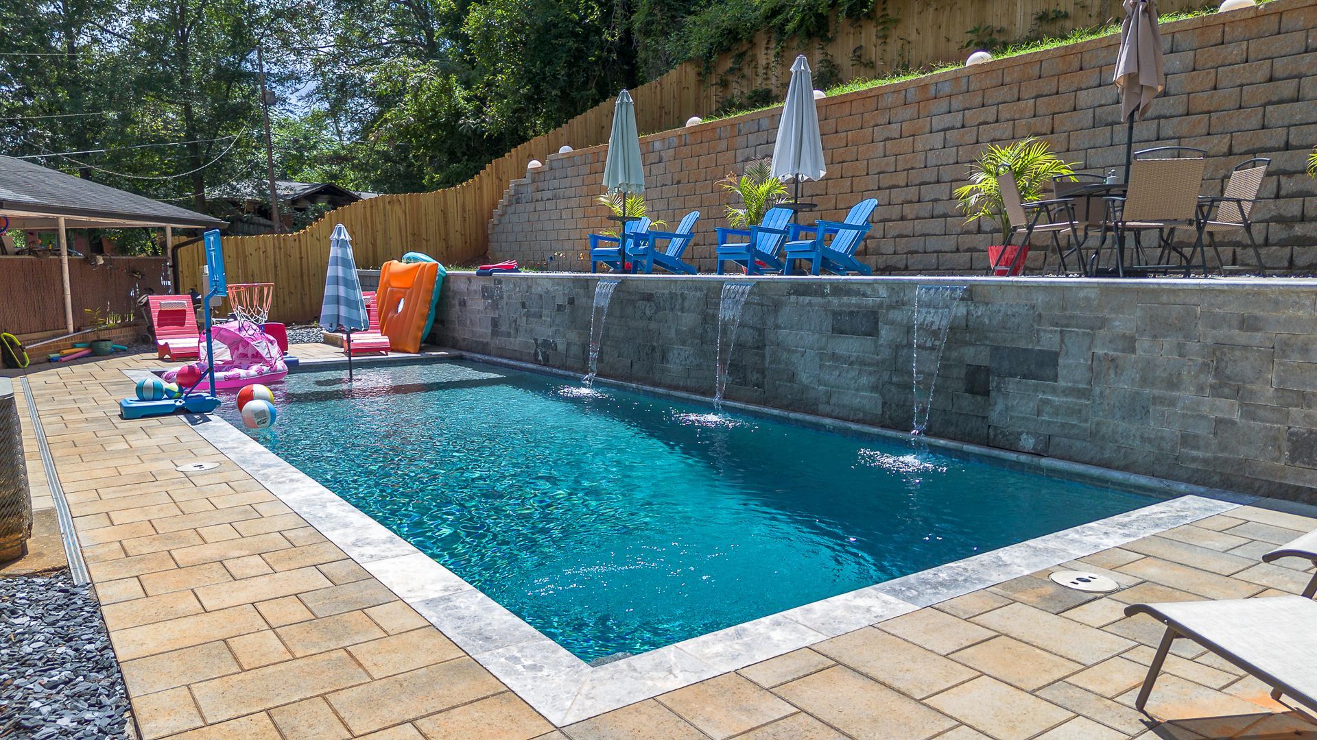 Backyard pool with water fountains and lounge chairs, against a retaining wall, sunny day.