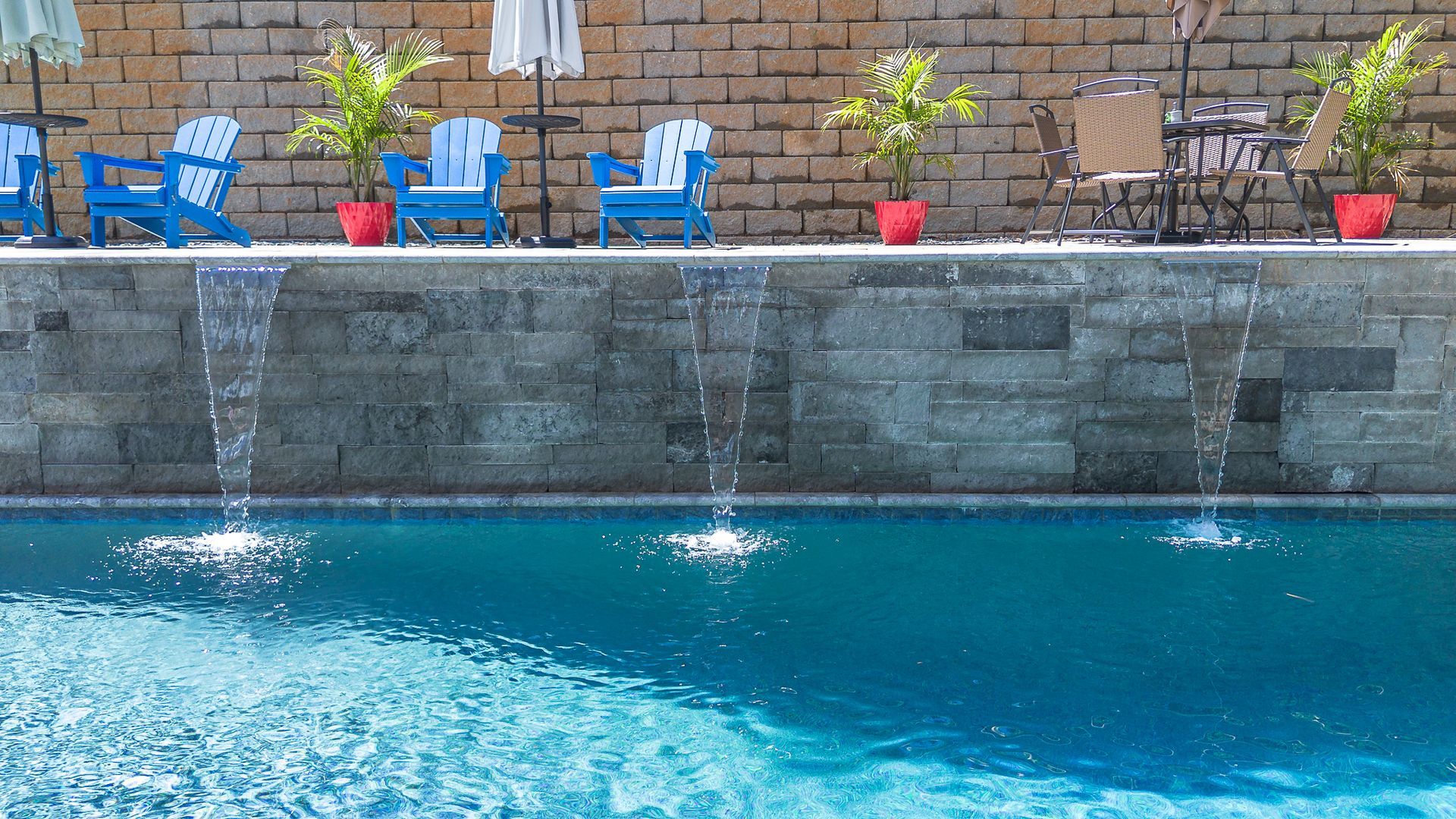 Pool with blue water, stone wall with water features, blue chairs, and small tables.