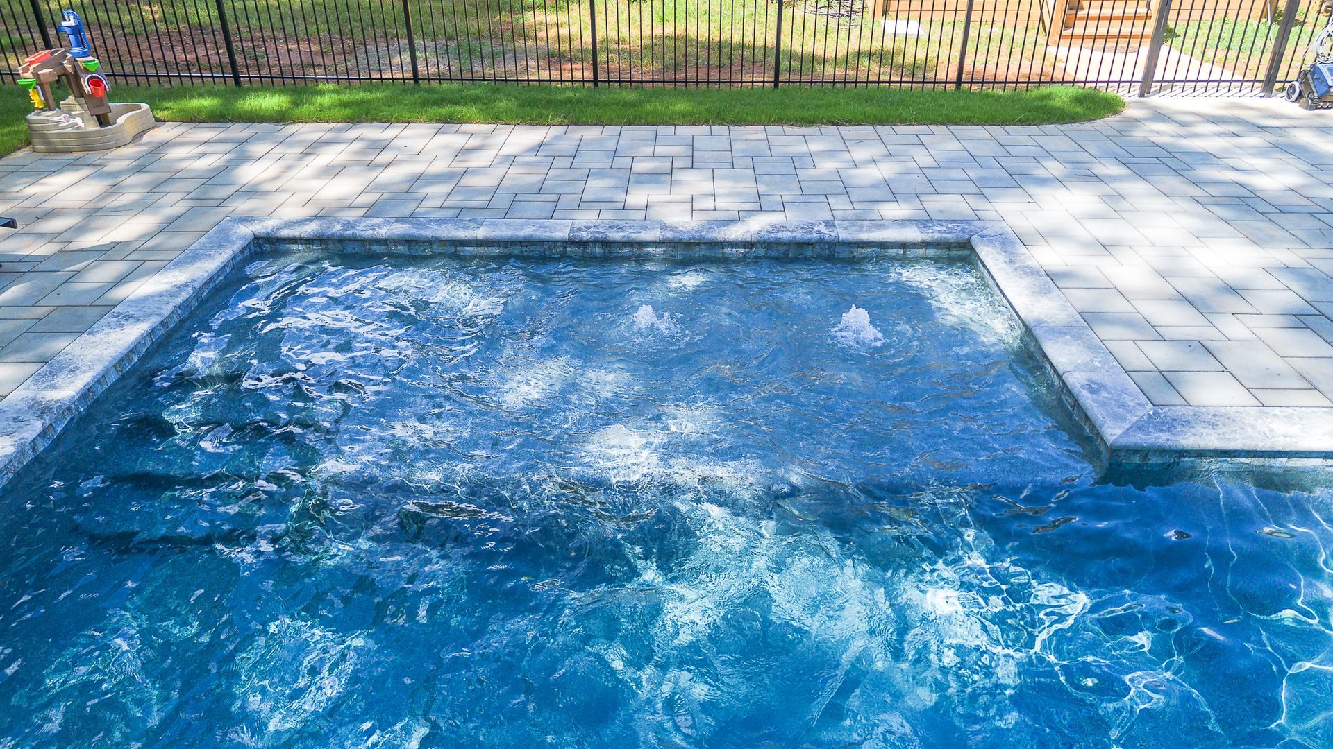 A pool with blue water and bubbling jets, surrounded by stone pavers and a fence.