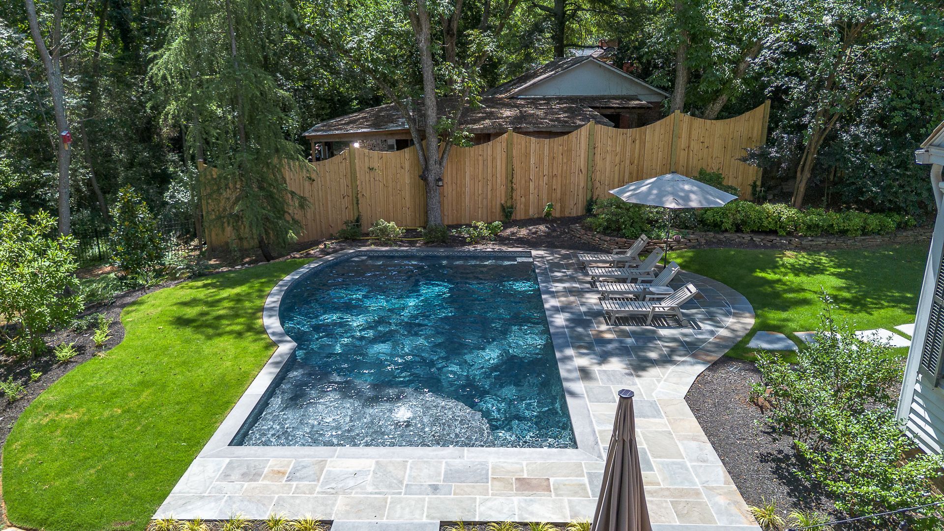 Backyard with a pool, lounge chairs, and a thatched fence surrounded by trees.
