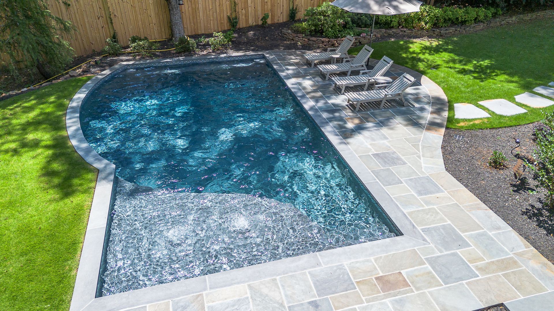 A small rectangular pool with blue water, surrounded by stone patio and grass, with lounge chairs.