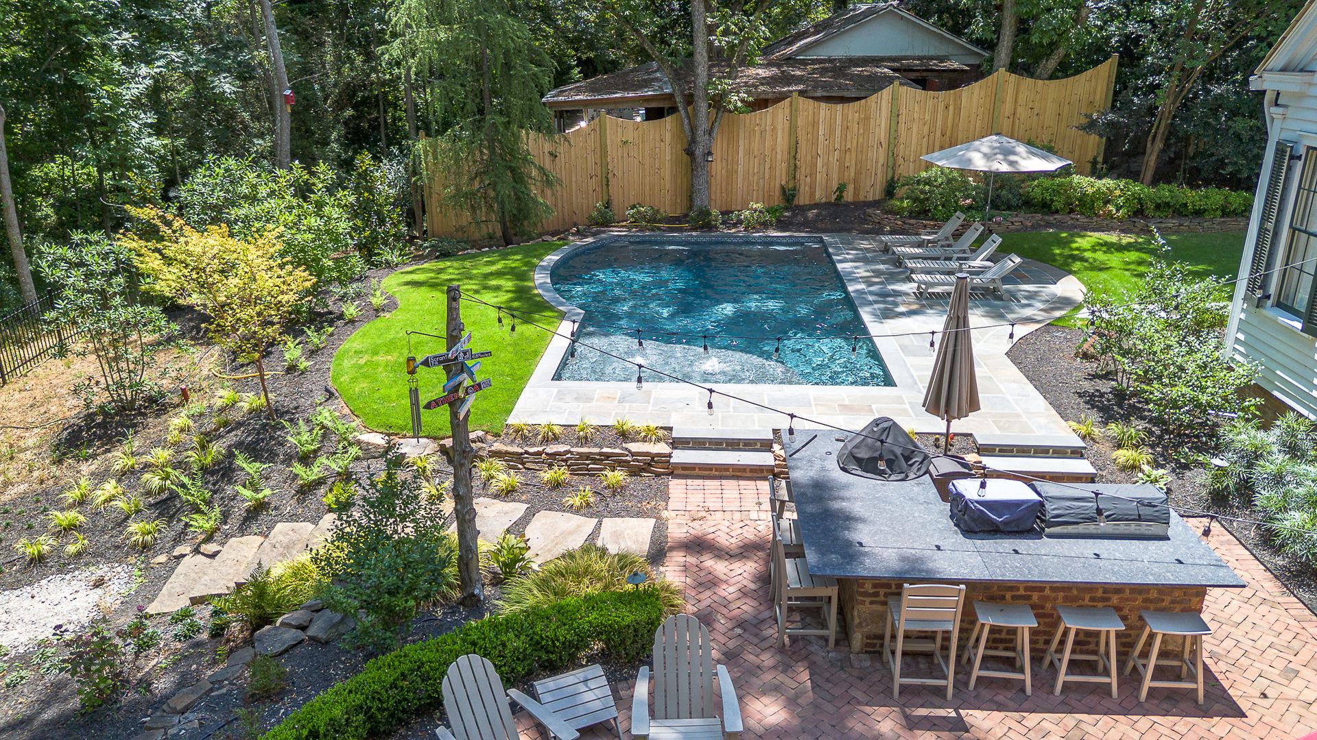 Backyard with a pool, outdoor kitchen, and seating. Green grass and trees surround the area.