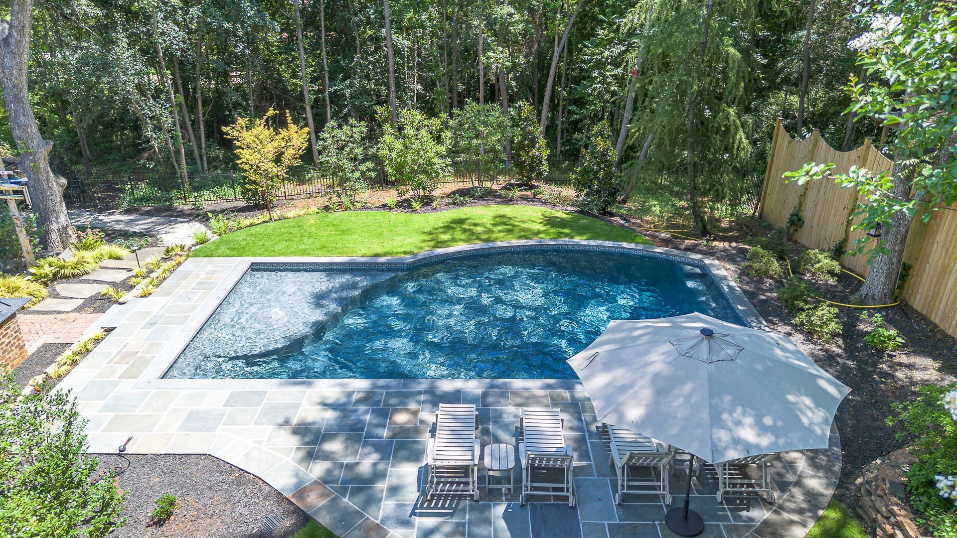 Rectangular pool with blue water, surrounded by stone patio, lounge chairs, and a large umbrella.