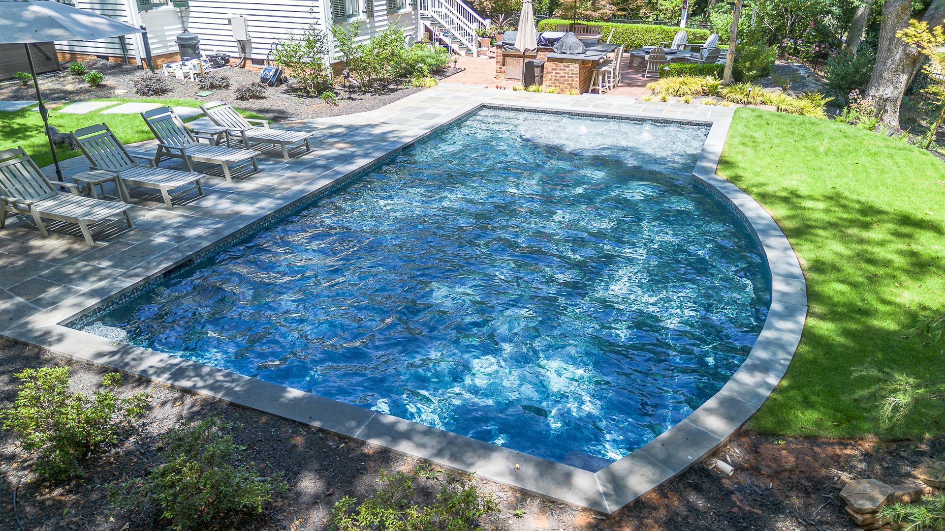 Rectangular pool with curved end, surrounded by stone, lawn, and lounge chairs; bright blue water.