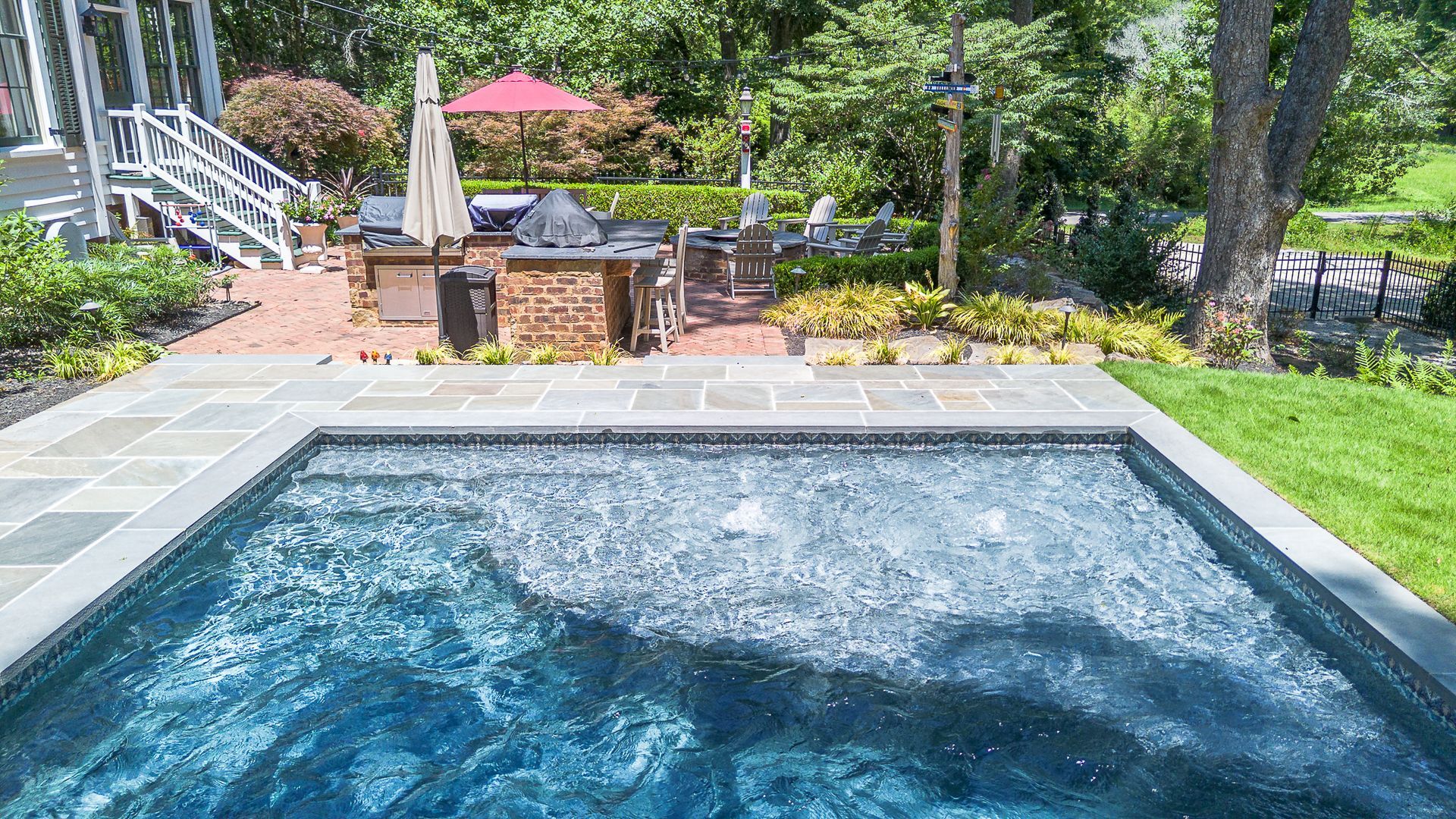 Swimming pool with stone border, overlooking a patio with a grill and seating, lush landscaping.