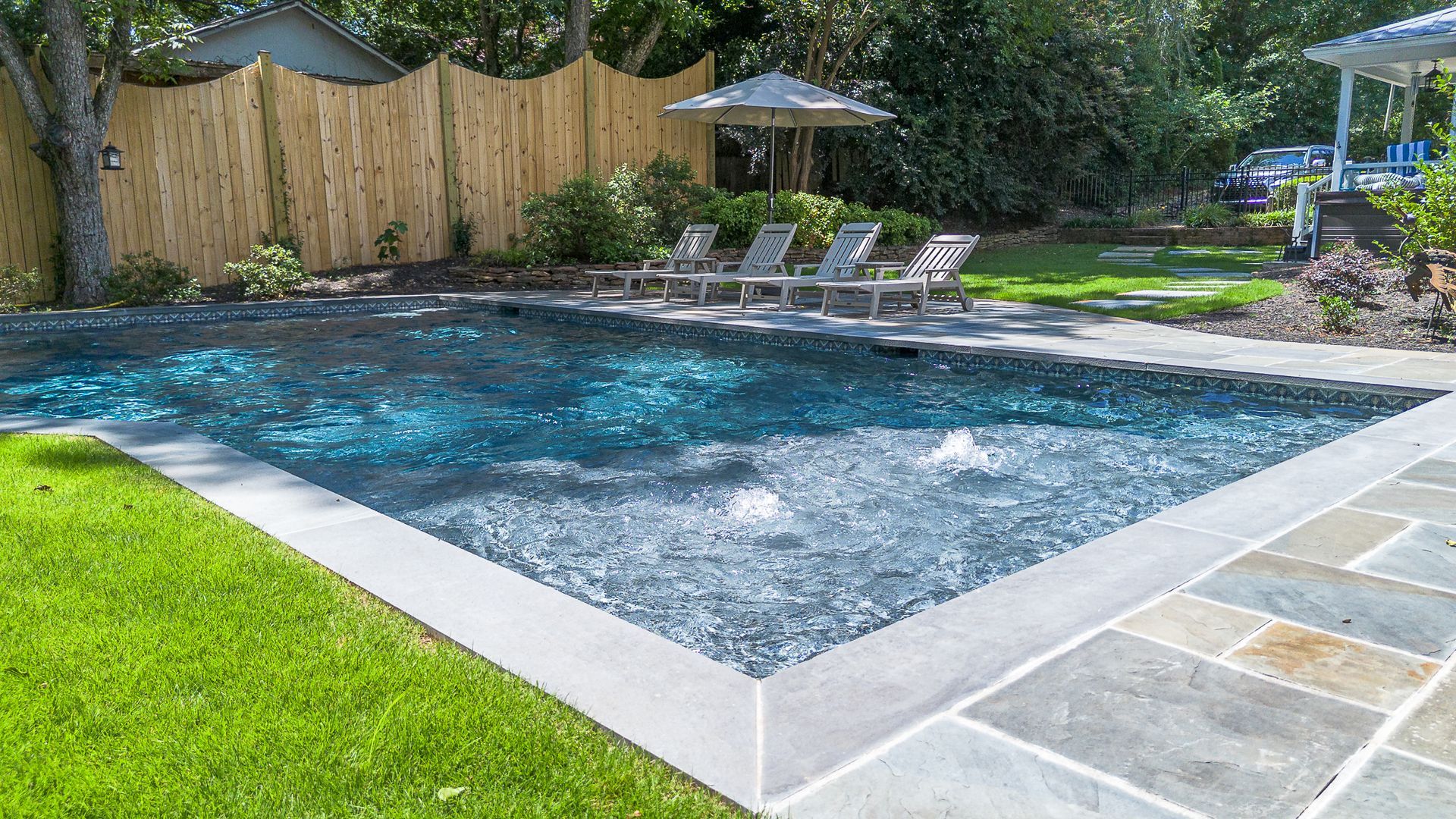 Swimming pool with blue water, surrounded by stone patio and green grass. Bamboo fence in background.