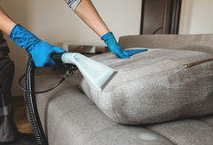 Person cleaning a beige couch with a handheld vacuum and blue gloves