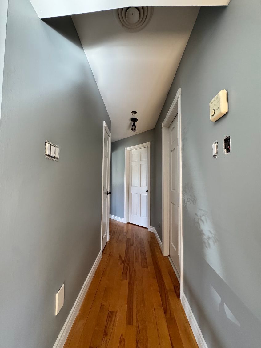 Couloir étroit aux murs gris, au sol en bois, aux portes et au plafond blancs, et équipé d'un thermostat.