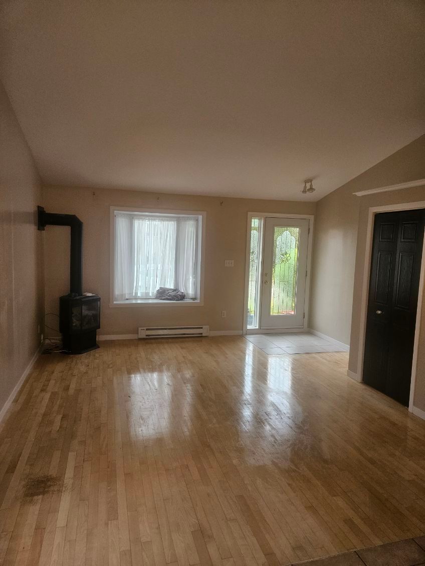 Salon vide avec parquet, poêle à bois, fenêtre et porte donnant sur l'extérieur.