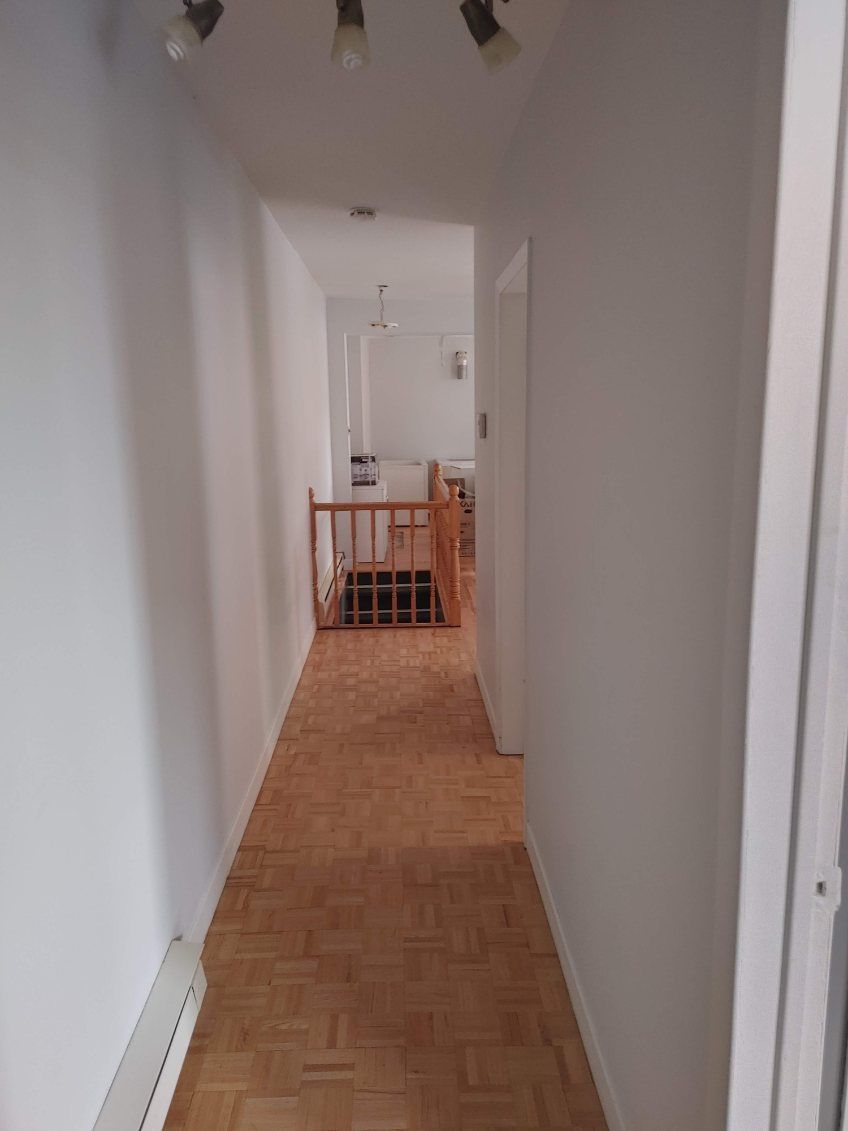 Long couloir aux murs blancs et au sol en bois, menant à un escalier avec une rampe en bois.