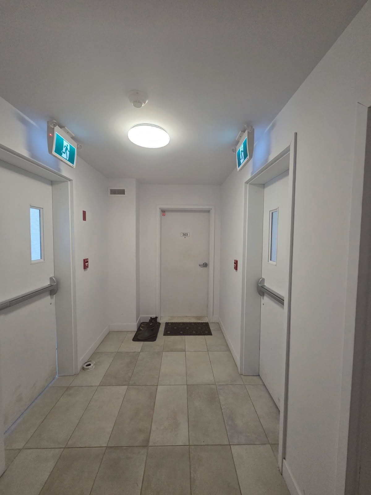 Couloir avec deux portes, panneaux de sortie de secours, éclairage au plafond et sol carrelé.