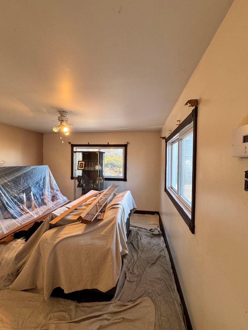 Chambre en rénovation avec meubles recouverts, encadrements de fenêtres sombres et parquet.