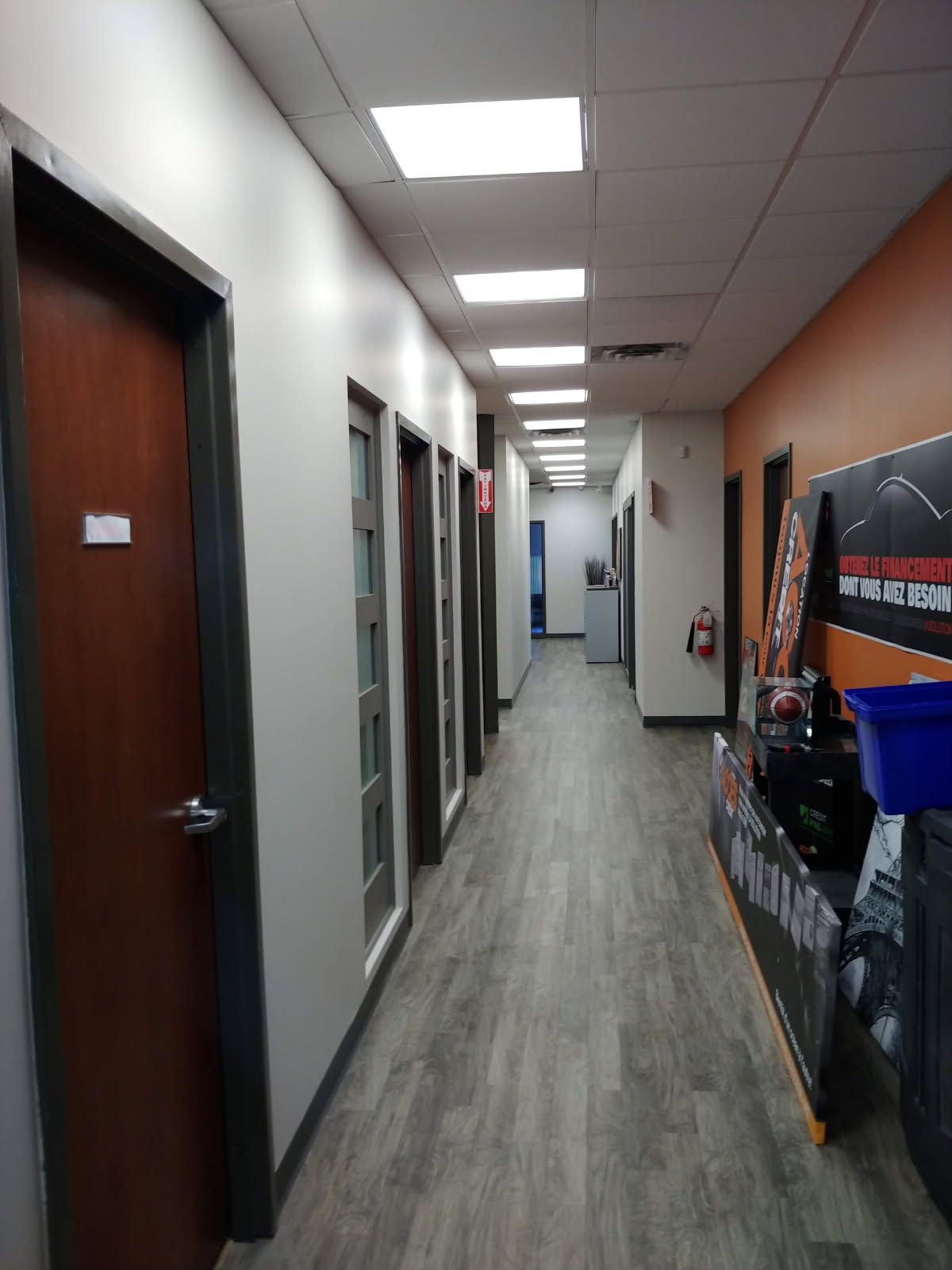 Couloir avec plusieurs portes de bureaux fermées, sol gris, murs blancs et orange, et éclairage zénithal.