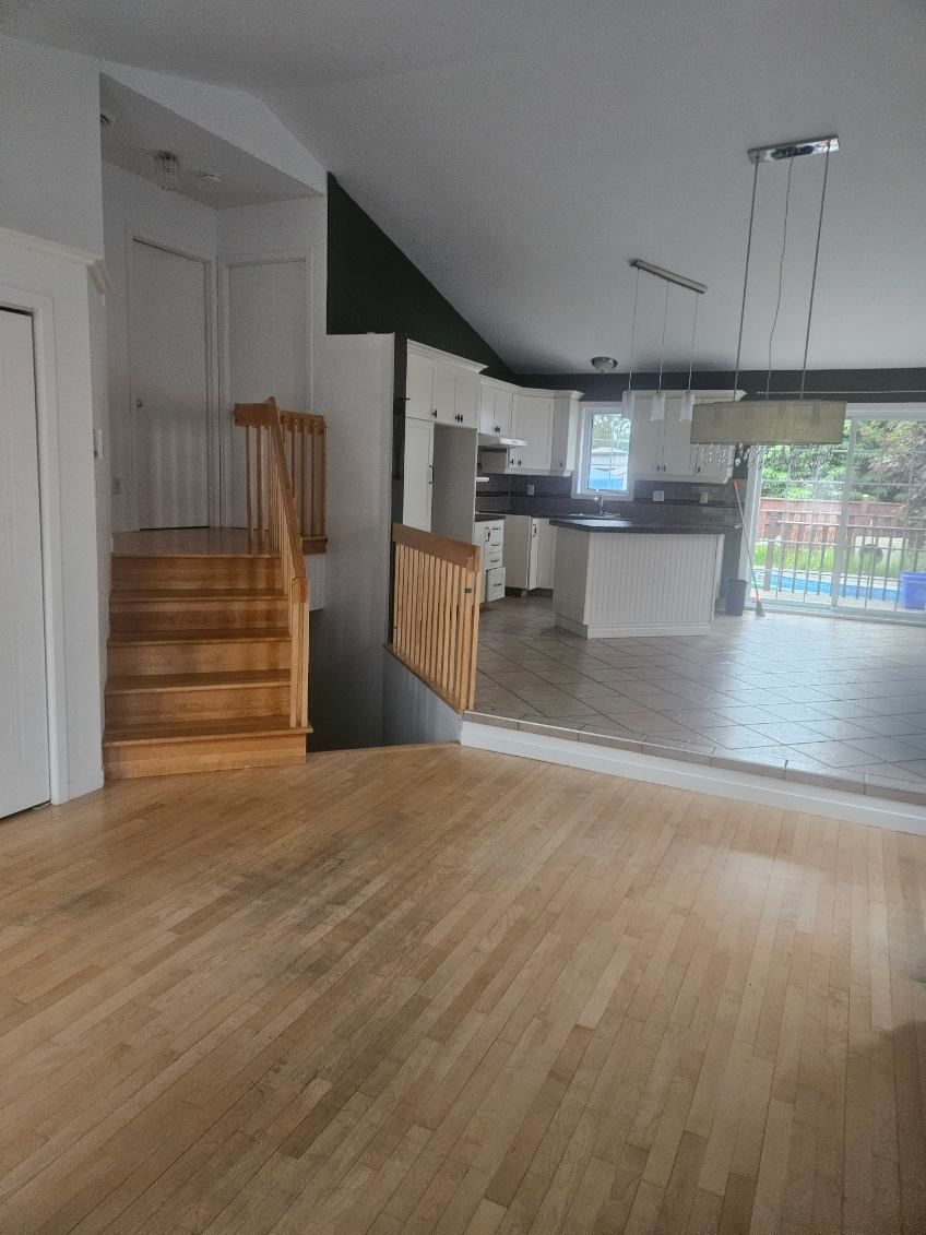 Escalier en bois menant à l'étage, cuisine ouverte en arrière-plan avec placards blancs et parquet.