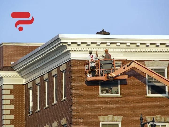 Deux ouvriers dans une nacelle peignent les boiseries blanches d'un immeuble en briques.