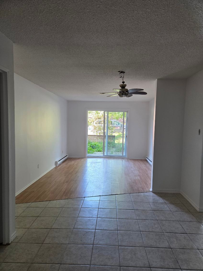 Pièce vide avec sol carrelé, se prolongeant par un parquet. Des portes coulissantes en verre donnent sur l'extérieur.