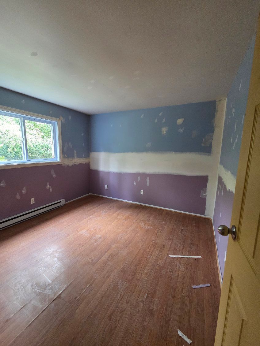 Pièce vide, murs peints en violet et bleu, fenêtre, parquet, porte à droite.