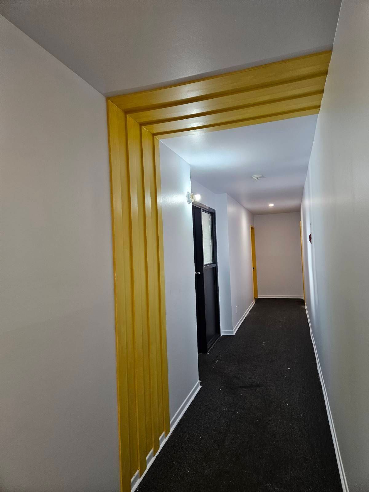 Couloir étroit aux murs gris, moquette noire et encadrement de porte jaune.