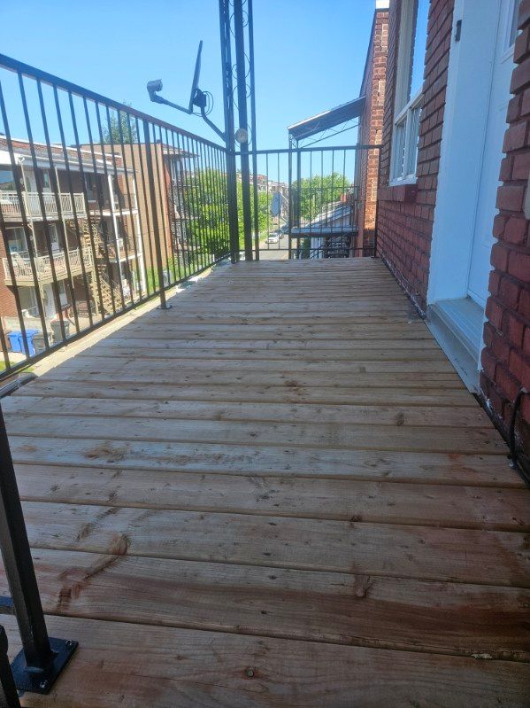Terrasse en bois sur un bâtiment en briques avec rambarde noire. Journée ensoleillée.