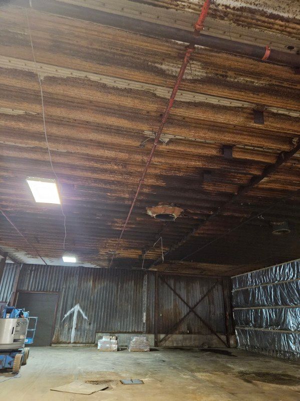 Intérieur d'entrepôt avec plafond et murs en tôle ondulée rouillée. Luminaires, panneau fléché et équipements visibles.