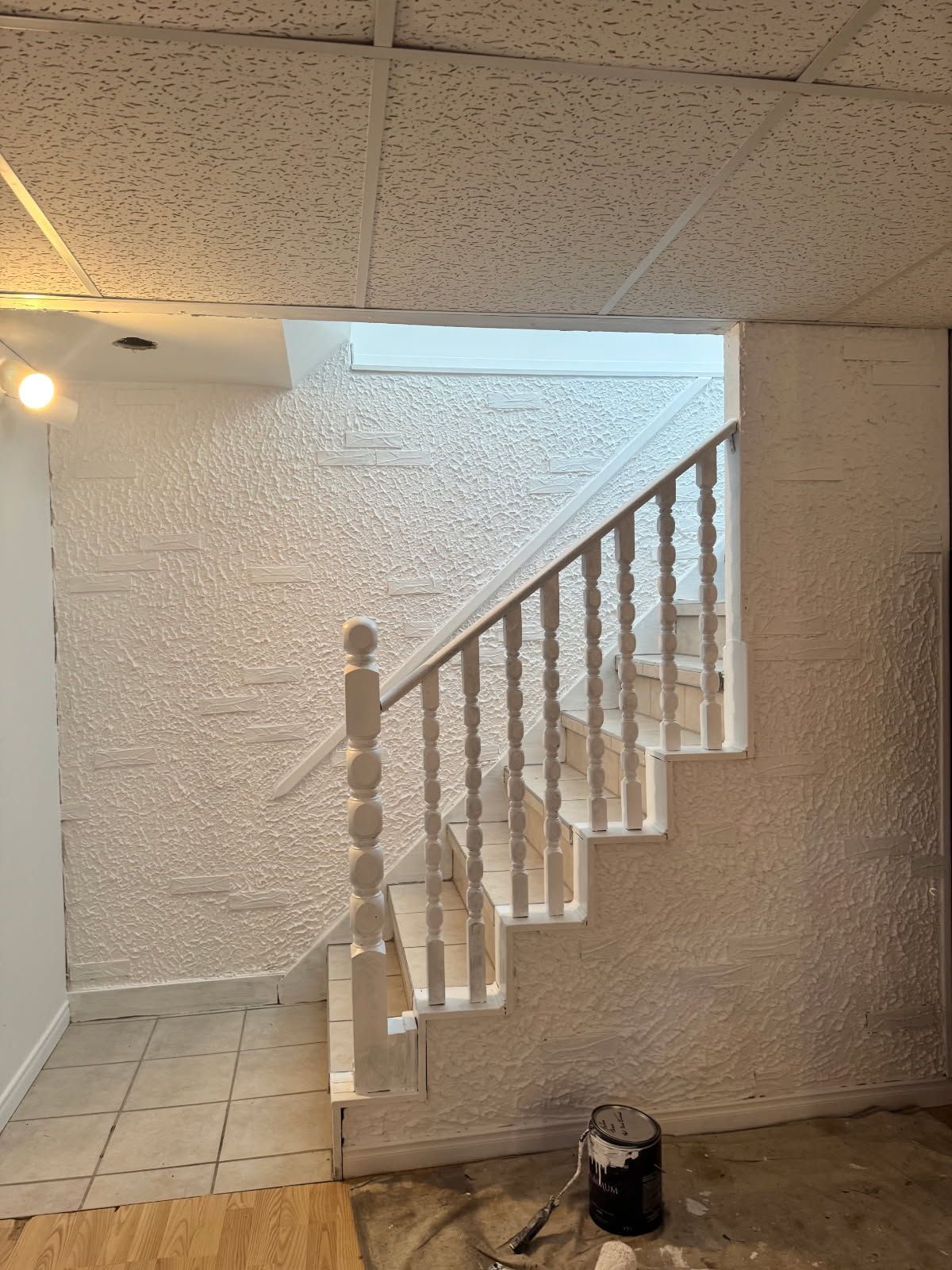 Escalier menant à l'étage, balustres blancs, murs blancs texturés, faux plafond, pot de peinture au sol.
