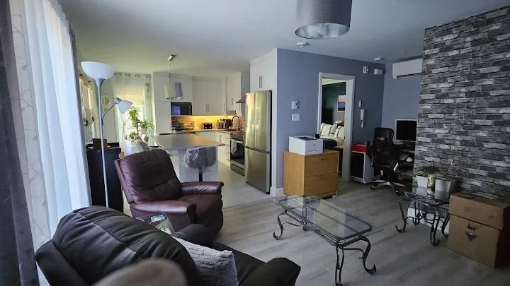 Salon avec cuisine ouverte, meubles en cuir marron, table basse en verre et mur en briques apparentes.