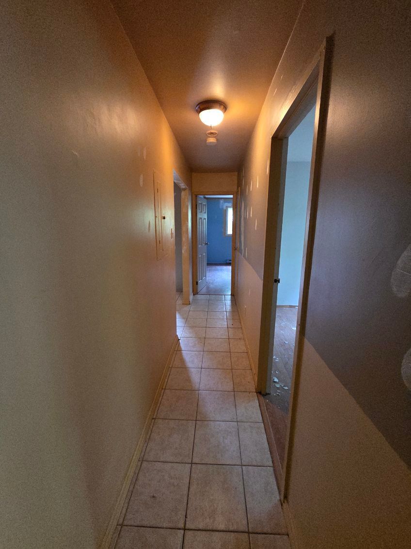 Couloir étroit aux murs beiges, au sol carrelé et aux portes sur la droite. Un plafonnier éclaire l'espace.